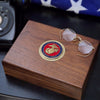 Wooden box with Marine Corps emblem and sunglasses on a dark surface with an American flag in the background