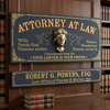A decorative attorney sign set displayed on a dark wood wall in front of bookshelves. The set includes a large arched sign reading 'ATTORNEY AT LAW' with a blindfolded bust in the center and a smaller rectangular sign below reading 'ROBERT G.